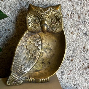 May include: A brass owl-shaped tray featuring intricate engraved feather details on the wings and head. The central body of the owl forms a smooth, concave basin with a weathered patina, while the head displays prominent circular eyes.