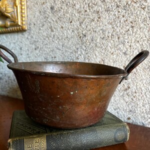 May include: A vintage copper bowl with two small handles. The bowl has a rounded shape and a rich, reddish-brown patina. It sits atop a weathered, green-covered book. The background features a textured wall and a glimpse of a gold-framed object.