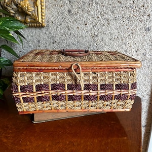 May include: A rectangular wicker basket with a hinged lid and a leather handle. The basket features a woven design with alternating bands of natural and burgundy-colored wicker. It sits on a wooden surface.