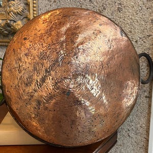 May include: A round, hammered copper pan featuring a weathered, mottled patina with dark spots and surface texture. The pan has two dark, circular metal handles attached to opposite sides, extending outward from the main copper body.