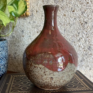 May include: A ceramic vase with a narrow neck and a rounded body. The upper portion features a glossy, deep red glaze, while the lower section has a textured, speckled beige and brown finish. The vase has a smooth, reflective surface.