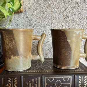May include: Two ceramic mugs with a two-tone glaze featuring a dark brown textured base and a lighter tan upper section. Each mug has a distinctively shaped, angular handle. The mugs are cylindrical with a smooth rim and a visible base ring.