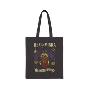 May include: Black tote bag with a graphic design featuring the text "HEX LA MIGRA" above an illustration of hands, a heart, and the word "ICE". The phrase "CURSES FOR CAGES" is printed below the illustration. The bag has two black handles.