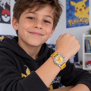 Può includere: Un orologio da polso analogico rotondo con quadrante giallo, illustrazione del personaggio Pikachu, numeri neri e cassa in metallo argentato. L'orologio presenta un cinturino in tessuto giallo brillante con motivo ripetuto e fibbia in metallo.