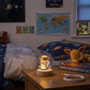 May include: A clear glass sphere night light featuring an internal 3D laser-etched Pikachu figure, resting on a circular light-up wooden base. The lamp is placed on a wooden surface next to a brown plush toy and two stacked books with text.