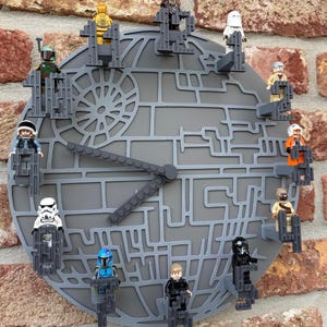 Könnte beinhalten: Eine kreisförmige graue Wanduhr mit einem Todesstern-Design und erhabenen Gittermustern. Zwölf kleine Kunststofffiguren sind an jeder Stundenposition auf dem Zifferblatt befestigt und dienen als Markierungen. Zwei lange graue Uhrzeiger sind mittig.
