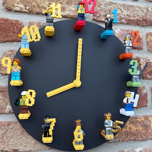 Könnte beinhalten: Eine schwarze runde Uhr mit gelben Zeigern und Lego-Figuren, die die Zahlen darstellen. Die Uhr ist an einer Ziegelmauer befestigt. Die Lego-Figuren sind in verschiedenen Farben gehalten und stellen verschiedene Charaktere dar.