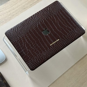 Può includere: Una custodia marrone per laptop con una texture a pelle di coccodrillo, con il logo Apple e la scritta "HEAVEN MAYHEM" in oro. La custodia è su un supporto in acrilico trasparente, con un mouse bianco e un portafoglio marrone con un logo visibile.