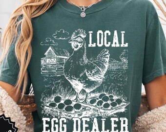 May include: Teal-colored t-shirt with a white graphic of a hen, eggs, and a barn, with the text "LOCAL EGG DEALER." The shirt also has the words "COMFORT COLORS" and "FAST SHIPPING" on it.