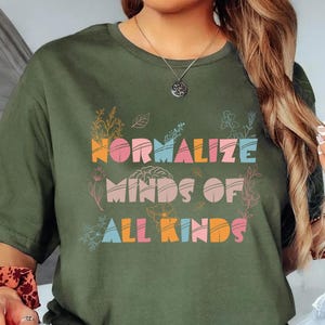 May include: Olive green t-shirt with the text "Normalize Minds of All Kinds" in colorful, stylized lettering. The design includes floral and botanical accents. The shirt is a casual, comfortable style.