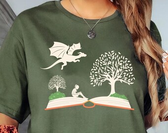 May include: Olive green t-shirt featuring a whimsical illustration of a dragon, trees, and a person reading on an open book. The design is in white, creating a striking contrast against the shirt's color. The shirt has short sleeves and a crew neck.