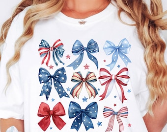 May include: White t-shirt featuring a pattern of watercolor bows in red, white, and blue. The bows are decorated with stars. The word "White" is printed in the lower right corner of the shirt.