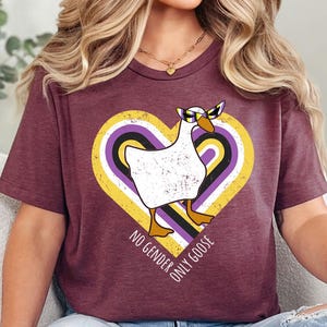 May include: Burgundy t-shirt featuring a white goose wearing sunglasses inside a heart-shaped design with non-binary flag colors. The text "NO GENDER ONLY GOOSE" is printed around the heart. The shirt is worn by a person.