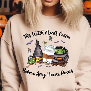 May include: Beige sweatshirt with a Halloween-themed graphic. The graphic includes a witch's hat, a coffee cup, a cauldron, and the text "This Witch Needs Coffee Before Any Hocus Pocus". Bats and a witch on a broom are also featured.