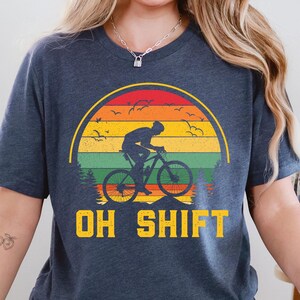 May include: Dark blue t-shirt featuring a cyclist silhouetted against a sunset design with red, orange, yellow, and green stripes. The text "OH SHIFT" is printed in yellow below the graphic. The shirt is a casual wear item.