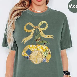 May include: A moss green short-sleeve t-shirt featuring a graphic print of two lemons tied with a yellow ribbon. The lemons are depicted with a patchwork-style pattern in shades of yellow, white, and blue, accented with small buttons and leaves.
