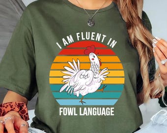 May include: Olive green t-shirt with a graphic of a white chicken and the text "I AM FLUENT IN FOWL LANGUAGE" in white. The chicken is set against a colorful sunset design. The shirt is a casual, everyday wear item.