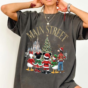 May include: A charcoal gray short-sleeve t-shirt featuring a graphic print of cartoon characters in holiday attire standing before a Christmas tree and a castle. The text MAIN STREET is printed in gold above the characters in a curved font.