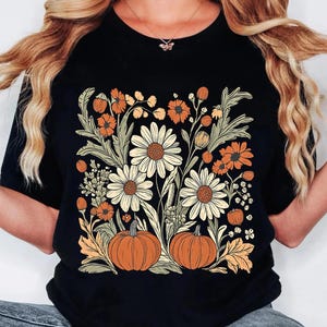 May include: Black t-shirt featuring a floral design with daisies, orange flowers, and pumpkins. The design includes green leaves and stems, creating a fall or autumn theme. The shirt is a classic crew neck style.