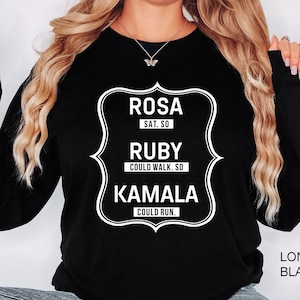 May include: Black long-sleeve sweatshirt with white text. The text reads "ROSA SAT. SO, RUBY COULD WALK. SO, KAMALA COULD RUN." The sweatshirt has a crew neck and a relaxed fit. The text is inside a white decorative frame.