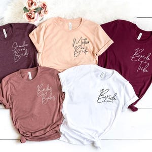 May include: Five t-shirts in various colors: burgundy, peach, mauve, and white. Each shirt has a different script text: "Grandma of the Bride", "Mother of the Bride", "Bride Tribe", "Brides Babes", and "Bride".