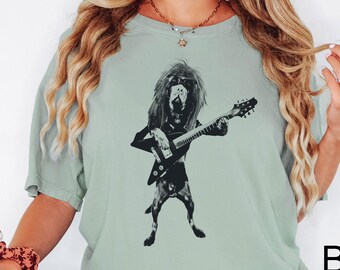 May include: A light green t-shirt featuring a black and white illustration of a dog playing an electric guitar. The dog has long hair and is wearing a jacket. The word "Bay" is printed in black on the bottom right.