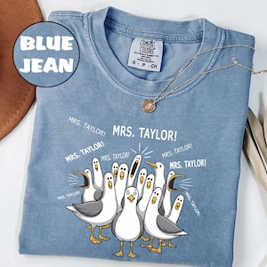 May include: A blue jean colored t-shirt featuring a graphic of multiple seagulls with open beaks and the repeated white text "MRS. TAYLOR!". The shirt has a Comfort Colors brand label at the neckline and a gold pendant necklace resting on the fabric.