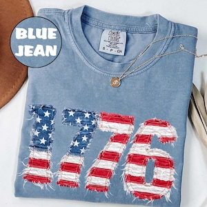 May include: A blue jean colored t-shirt featuring the number 1776 appliqued in a frayed American flag pattern. The shirt has a crew neckline and a Comfort Colors brand label at the collar. A gold-toned necklace with a small circular pendant rests on the fabric.