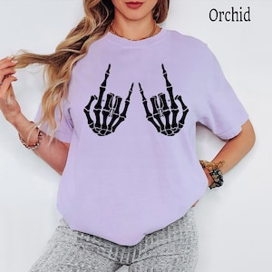 Puede incluir: Una camiseta de manga corta de color morado claro con un estampado gr&aacute;fico negro de dos manos de esqueleto haciendo el gesto de rock and roll. El texto "Orchid" est&aacute; impreso en la esquina superior derecha de la prenda de cuello redondo.