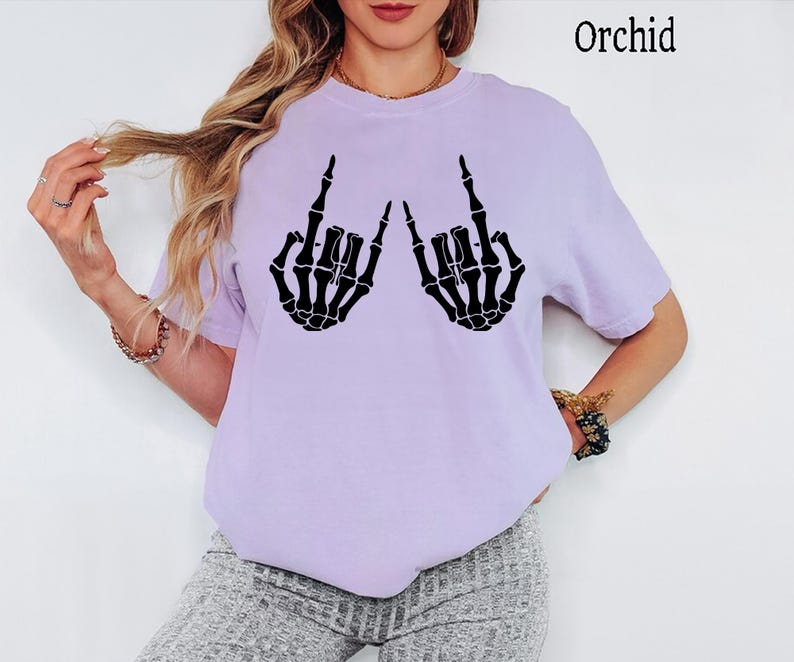 Puede incluir: Una camiseta de manga corta de color morado claro con un estampado gr&aacute;fico negro de dos manos de esqueleto haciendo el gesto de rock and roll. El texto "Orchid" est&aacute; impreso en la esquina superior derecha de la prenda de cuello redondo.