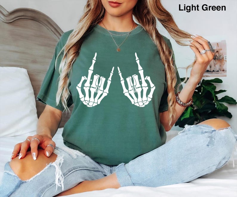 Puede incluir: Una camiseta de color verde claro con un gr&aacute;fico blanco de dos manos de esqueleto haciendo el gesto de rock and roll. La prenda tiene cuello redondo y mangas cortas. El texto Light Green aparece en la esquina superior derecha de la imagen.