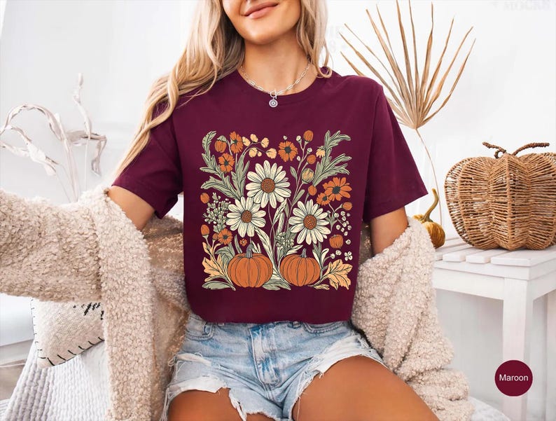 K&ouml;nnte beinhalten: Burgunderfarbenes T-Shirt mit floralem Design mit G&auml;nsebl&uuml;mchen, orangefarbenen Blumen und K&uuml;rbissen. Die Grafik ist mittig auf der Vorderseite. Das kurz&auml;rmelige T-Shirt ist aus weichem Material. Es wird mit Jeansshorts getragen.
