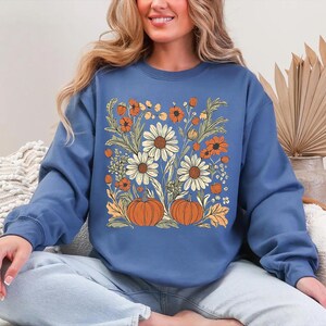 K&ouml;nnte beinhalten: Ein blaues Sweatshirt mit einem Blumendesign mit G&auml;nsebl&uuml;mchen, orangefarbenen Blumen und K&uuml;rbissen. Das Design ist auf der Vorderseite zentriert. Das Sweatshirt hat lange &Auml;rmel und einen Rundhalsausschnitt.