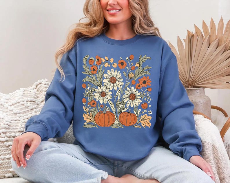 K&ouml;nnte beinhalten: Ein blaues Sweatshirt mit einem Blumendesign mit G&auml;nsebl&uuml;mchen, orangefarbenen Blumen und K&uuml;rbissen. Das Design ist auf der Vorderseite zentriert. Das Sweatshirt hat lange &Auml;rmel und einen Rundhalsausschnitt.