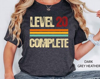May include: Dark grey heather t-shirt with the text "LEVEL 20 COMPLETE" in a distressed font. The number 20 is red, and the text is above three horizontal stripes in orange, yellow, and teal.