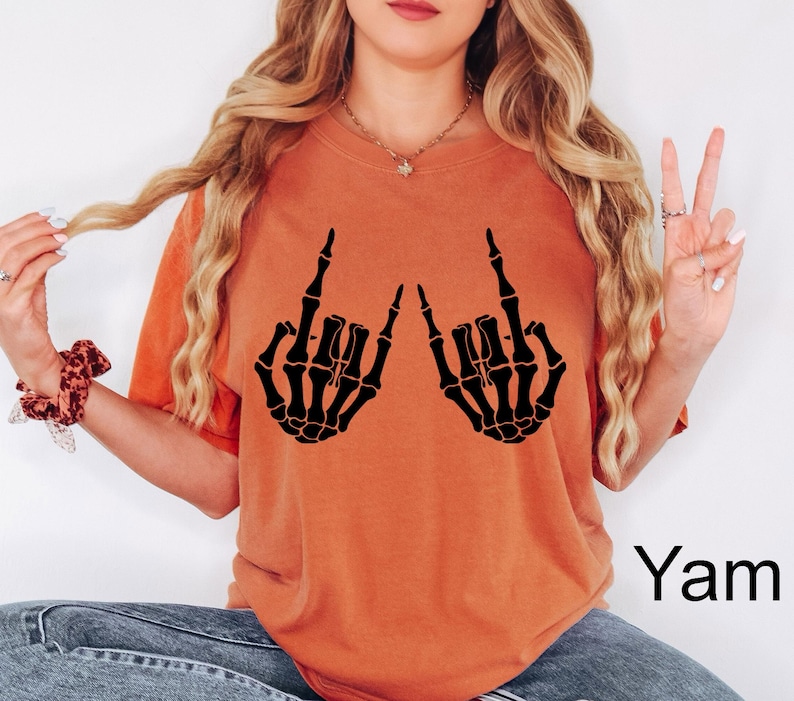 Puede incluir: Una camiseta de manga corta color naranja quemado con un estampado gr&aacute;fico negro de dos manos de esqueleto haciendo el gesto de rock and roll. La camiseta tiene cuello redondo y un corte holgado. La palabra Yam est&aacute; impresa en negro abajo.