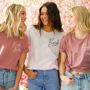 May include: Three women wearing t-shirts with bridal party-themed text. One woman wears a white shirt with "Bride" in script. The other two wear dusty rose shirts with "Bride Babes" and "Bride Tribe" in script. All are wearing blue jeans.