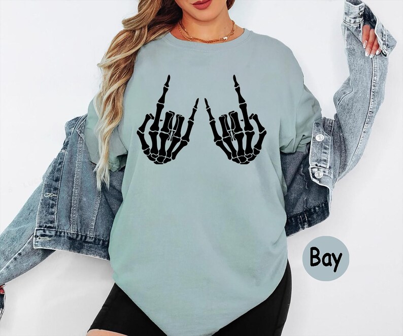 Puede incluir: Una camiseta de cuello redondo de color azul claro con un estampado gr&aacute;fico negro de dos manos de esqueleto haciendo un gesto de rock and roll. La camiseta se lleva bajo una chaqueta vaquera azul y pantalones cortos negros. La palabra Bay est&aacute; impresa en negro.