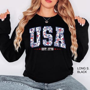 May include: Black long-sleeve sweatshirt with the word "USA" in a floral pattern, with the text "EST. 1776" below. The letters are white with a floral design of red and blue flowers.