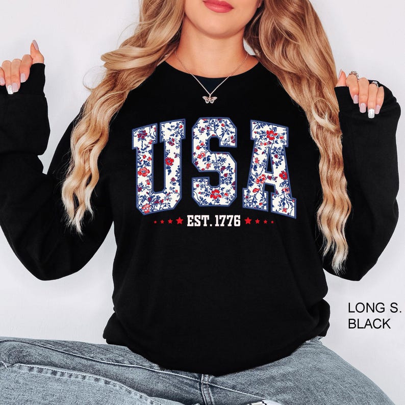 May include: Black long-sleeve sweatshirt with the word "USA" in a floral pattern, with the text "EST. 1776" below. The letters are white with a floral design of red and blue flowers.