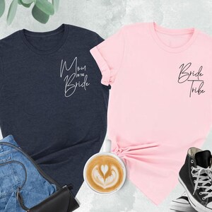 May include: Two t-shirts, one navy blue and one pink, with script text. The navy shirt says "Mom of the Bride," and the pink shirt says "Bride Tribe." The shirts are displayed with jeans, a black purse, a cup of coffee, and black high-top sneakers.