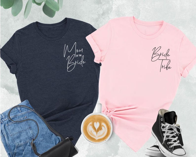 May include: Two t-shirts, one navy blue and one pink, with script text. The navy shirt says "Mom of the Bride," and the pink shirt says "Bride Tribe." The shirts are displayed with jeans, a black purse, a cup of coffee, and black high-top sneakers.