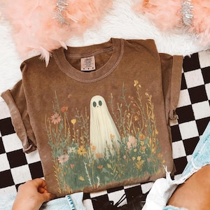 May include: Brown Comfort Colors t-shirt featuring a whimsical ghost design in a field of wildflowers. The ghost is white with black eyes and is surrounded by colorful flowers and green grass. The shirt has a relaxed fit and short sleeves.