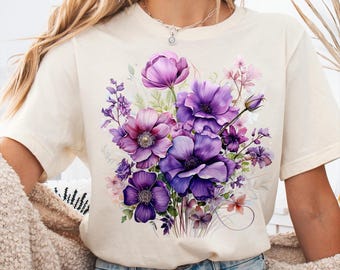 May include: Cream-colored t-shirt featuring a watercolor floral design in shades of purple, pink, and green. The flowers are clustered together, creating a vibrant and artistic display. The shirt has a relaxed fit.
