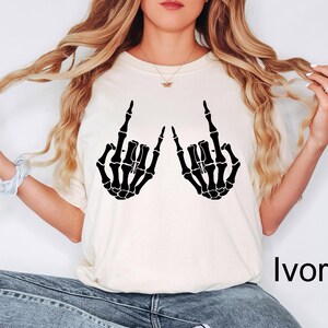 Puede incluir: Una camiseta de manga corta de color marfil con un estampado gr&aacute;fico negro de dos manos de esqueleto haciendo el signo de mano de rock and roll. La palabra Ivory est&aacute; impresa en negro en la esquina inferior derecha de la imagen.
