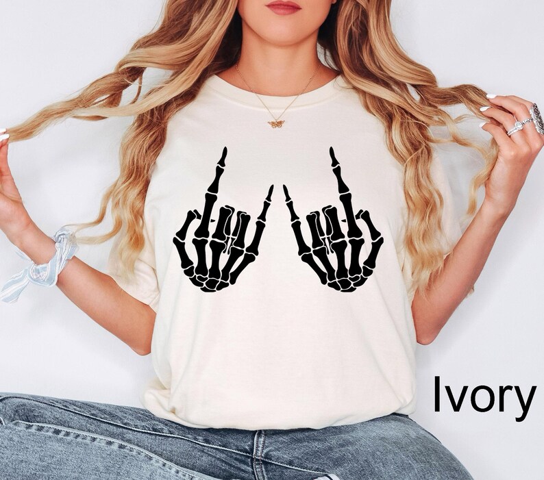 Puede incluir: Una camiseta de manga corta de color marfil con un estampado gr&aacute;fico negro de dos manos de esqueleto haciendo el signo de mano de rock and roll. La palabra Ivory est&aacute; impresa en negro en la esquina inferior derecha de la imagen.