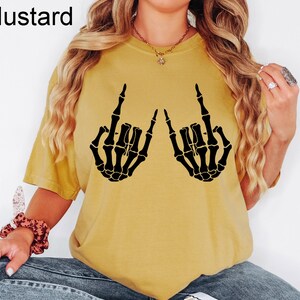 Puede incluir: Una camiseta de manga corta color amarillo mostaza con un estampado gr&aacute;fico negro de dos manos de esqueleto haciendo el signo de mano de rock and roll. La palabra Mustard est&aacute; impresa en la esquina superior izquierda de la imagen.