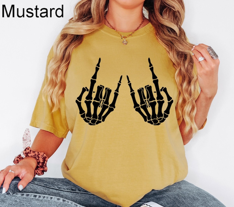 Puede incluir: Una camiseta de manga corta color amarillo mostaza con un estampado gr&aacute;fico negro de dos manos de esqueleto haciendo el signo de mano de rock and roll. La palabra Mustard est&aacute; impresa en la esquina superior izquierda de la imagen.