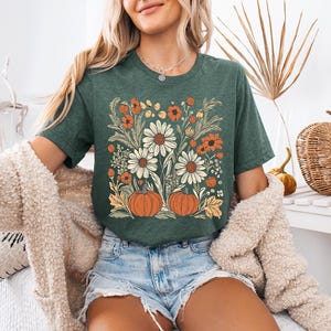 K&ouml;nnte beinhalten: Gr&uuml;nes T-Shirt mit einem Blumendesign mit wei&szlig;en G&auml;nsebl&uuml;mchen, orangefarbenen Blumen und zwei orangefarbenen K&uuml;rbissen. Das T-Shirt hat einen Rundhalsausschnitt und kurze &Auml;rmel. Das Thema ist Herbst.