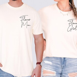 May include: Cream-colored t-shirts with the phrases "Flower Man" and "Flower Girl" in black script. The shirts are short-sleeved and appear to be made of a soft material. The shirts are worn by two people.